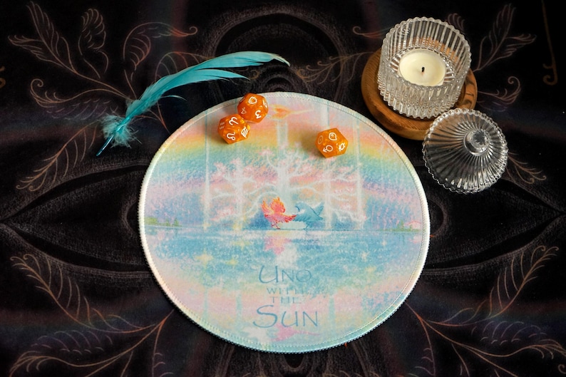 May include: A round, pastel-colored mousepad with the text "Uno with the Sun" and a whimsical illustration. Three orange dice and a blue feather are on the dark background. A glass candle holder and a small glass jar are also present.