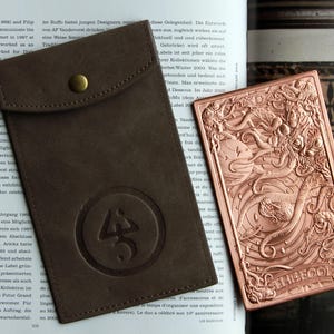 May include: A dark brown leather pouch with a brass snap button and a circular embossed logo, paired with a rectangular copper-toned metal plate featuring an intricate relief design of a sea creature and the word THE FOOL at the bottom.