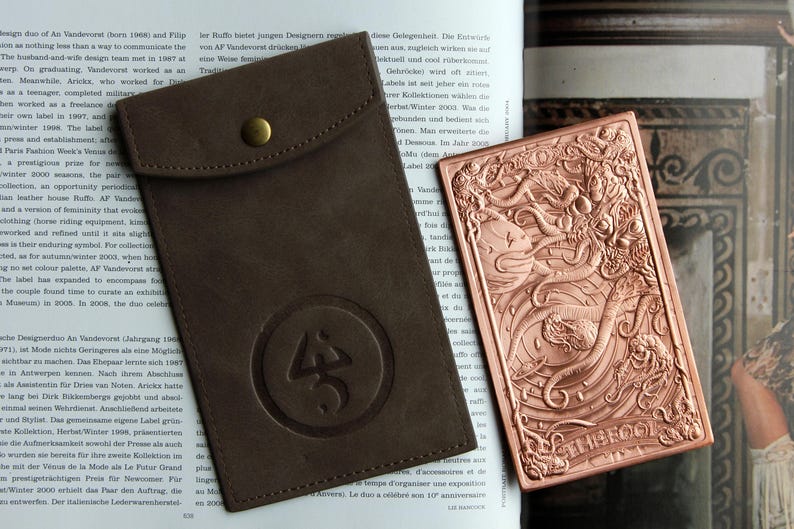 May include: A dark brown leather pouch with a brass snap button and a circular embossed logo, paired with a rectangular copper-toned metal plate featuring an intricate relief design of a sea creature and the word THE FOOL at the bottom.