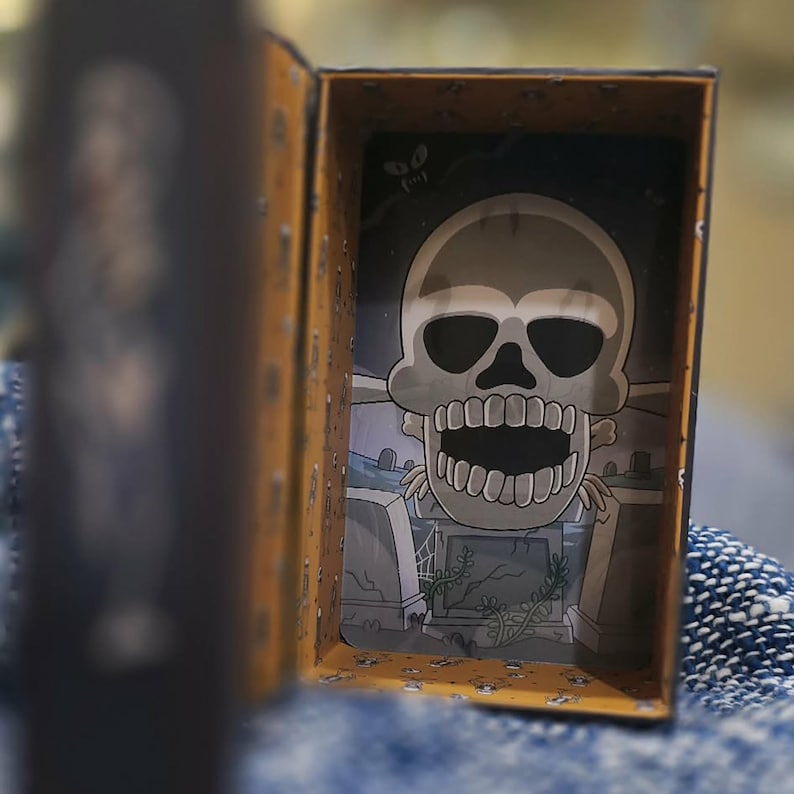 May include: An open, rectangular box with a skull illustration inside. The skull is gray with a wide open mouth, set against a graveyard scene. The box has a yellow-orange exterior and a blurred image of a person on the left.