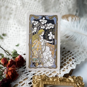 May include: A vintage-style tarot card with a gold frame and a woman in a white dress surrounded by flowers. The card features the word "Happiness" in gold lettering.