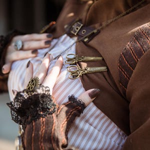 Puede incluir: Una chaqueta marrón con una camisa a rayas blancas y marrones debajo. La chaqueta tiene una cadena decorativa con un par de tijeras antiguas sujetas.