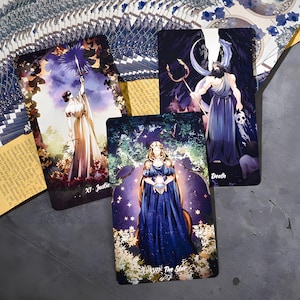 Puede incluir: Un mazo de cartas del tarot con ilustraciones de figuras y s&iacute;mbolos. Las cartas est&aacute;n dispuestas sobre una superficie oscura, con las cartas "Justicia", "Muerte" y "La Estrella" a la vista. El mazo tiene un tema floral y celestial.
