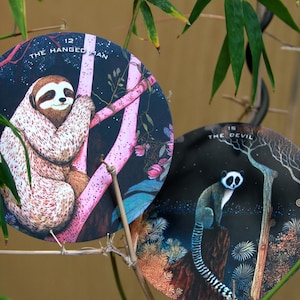 May include: Two round tarot cards featuring animal illustrations. One card shows a sloth labeled "The Hanged Man" and the other depicts a lemur labeled "The Devil". The cards are set against a dark background with pink and brown tree branches.