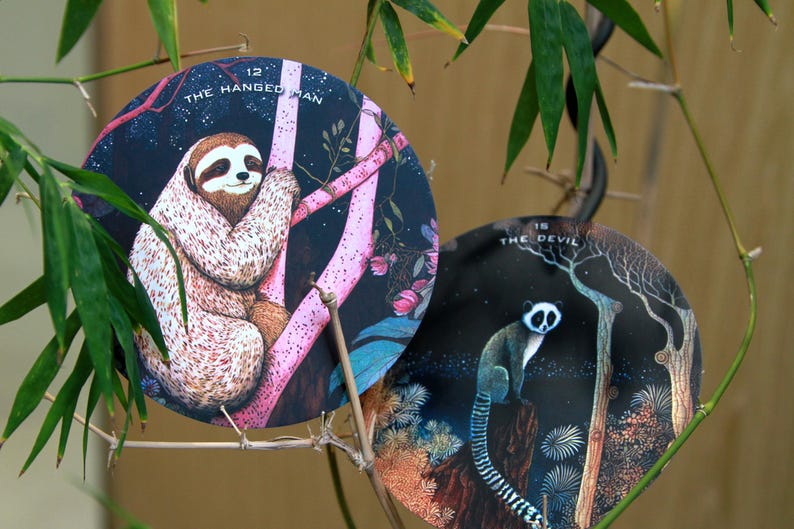 May include: Two round tarot cards featuring animal illustrations. One card shows a sloth labeled "The Hanged Man" and the other depicts a lemur labeled "The Devil". The cards are set against a dark background with pink and brown tree branches.