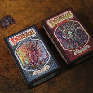 May include: Two rectangular card game boxes titled Call of the Pantheon Cthulhu Mythos Cards. The left box features a holographic blue and purple monster illustration, while the right box displays a red and purple monster with multiple eyes.