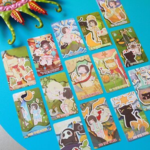 May include: A collection of colorful tarot cards featuring whimsical illustrations and text. Each card displays unique artwork with titles like "The Magician," "The Fool," and "The Star." The cards are arranged on a bright blue surface, with a decorative green and red object in the corner.