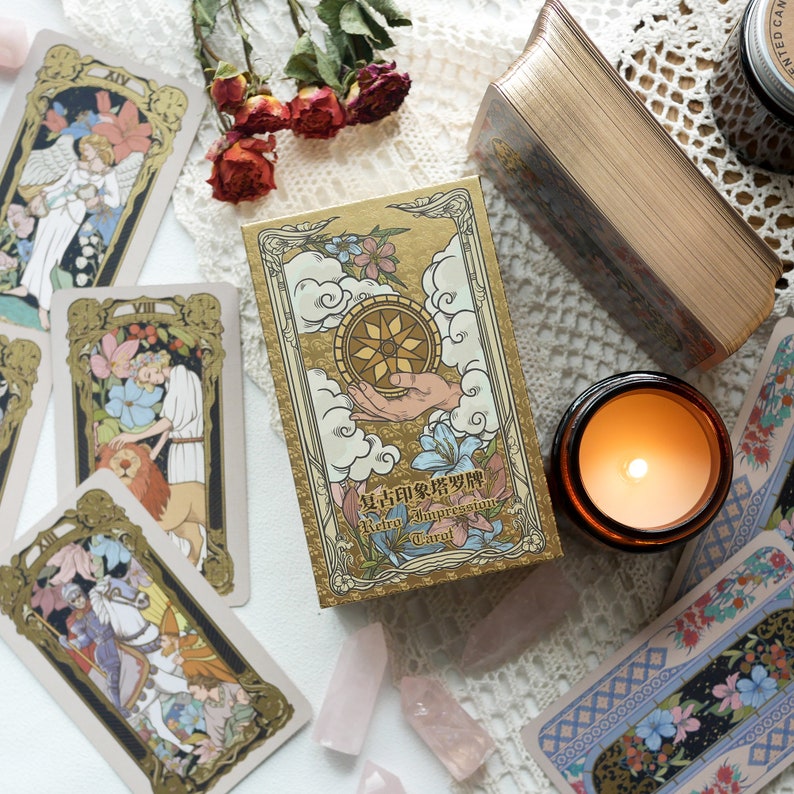 May include: A gold box with a tarot card deck inside. The box has a gold and white design with the text "Retro Impression Tarot" on the front. The box is surrounded by tarot cards, flowers, and crystals.