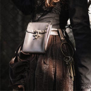 May include: A brown leather bag with a metal buckle and rivets. The bag is attached to a brown belt with a silver buckle. The bag is worn over a brown skirt with a black top.