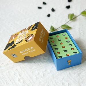 Puede incluir: Una caja amarilla con una baraja de tarot con tem&aacute;tica de gatos en su interior. La caja tiene caracteres chinos y las palabras "Miaow Tarot". La baraja est&aacute; en una caja azul con una carta verde que muestra un patr&oacute;n de flores blancas y tulipanes naranjas.