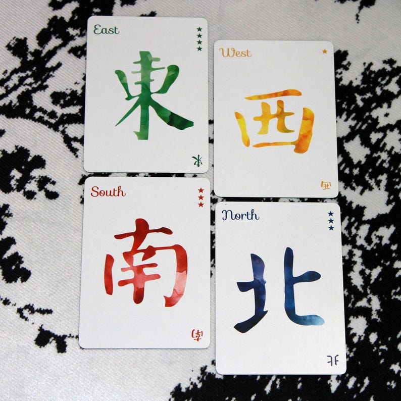 Pu&ograve; includere: Quattro carte bianche con calligrafia ad acquerello che rappresentano Est, Ovest, Sud e Nord. Ogni carta presenta un colore e un simbolo diversi. Le carte sono disposte su una superficie a motivi.