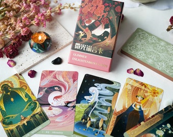 May include: A deck of Glimmer Enlightenment Cards with a box featuring a colorful illustration of a woman in a forest. The cards are spread out on a white surface with pink flowers, a candle, and other decorative elements.