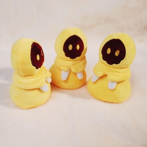 Op de afbeelding: Drie gele pluche figuren met donkerbruine gezichten en gele ogen. Elke figuur heeft een capuchon en een witte kraag. De knuffels zijn gemaakt van zacht materiaal.