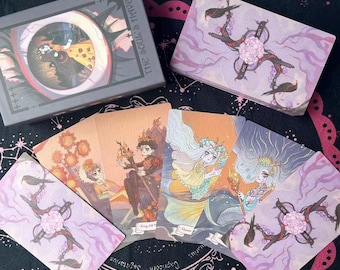 Può includere: Un mazzo di carte dei tarocchi con una scatola grigia con la scritta "THE SOUL'S HAVEN". Le carte presentano illustrazioni di figure, disegni floreali e i testi "King of Wands" e "Queen". Le carte sono su una superficie nera.