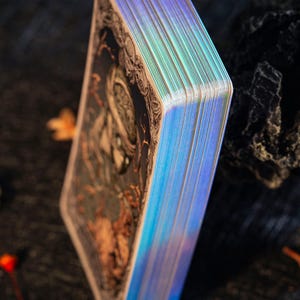May include: A deck of rectangular cards with dark, intricate illustrations on the back and reflective, iridescent holographic edges. The cards are stacked vertically, showcasing the metallic sheen of the sides against a dark, textured surface.