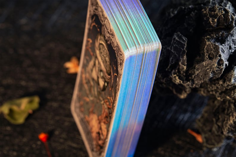 May include: A deck of rectangular cards with dark, intricate illustrations on the back and reflective, iridescent holographic edges. The cards are stacked vertically, showcasing the metallic sheen of the sides against a dark, textured surface.