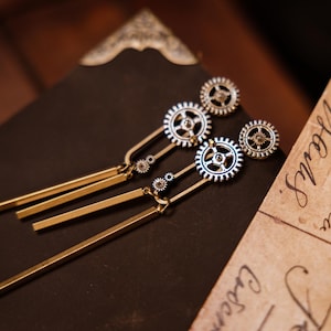 May include: A pair of gold and silver dangle earrings with a gear design. The earrings have long, thin, gold bars hanging from the gears.