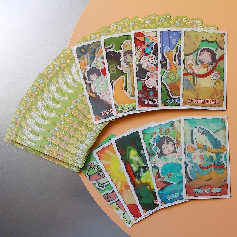 May include: A deck of tarot cards with whimsical illustrations. The cards feature colorful artwork with figures and symbols, including The Empress and Page of Cups. The card backs have a light green floral pattern. The cards are arranged in a fan shape.