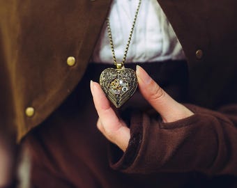 Puede incluir: Un medallón de corazón de oro con detalles intrincados y un mecanismo de relojería visible en su interior. El medallón cuelga de una cadena de oro.