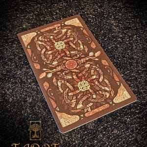 May include: A tarot card with a brown background and detailed Celtic knot patterns in brown, beige, and gold. The card has the text "Tarot The Forest Knows" at the bottom, with a tree logo.