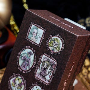May include: A brown rectangular tarot deck box featuring a dark damask pattern and multiple framed illustrations of stylized figures. The text "Wohstudios" and "Doll House tarot" is printed in white on the box surface.