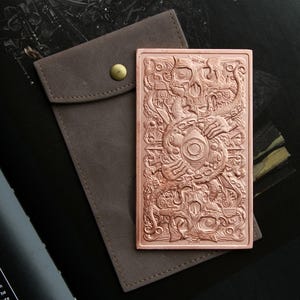 May include: A rectangular copper-colored metal plate featuring an intricate embossed design of a central eye surrounded by tentacles and skulls, accompanied by a brown fabric pouch with a brass-colored snap button closure.