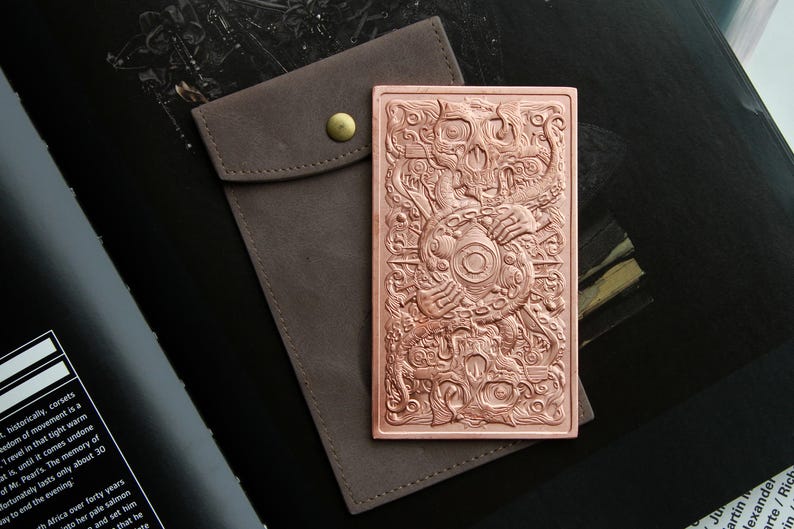 May include: A rectangular copper-colored metal plate featuring an intricate embossed design of a central eye surrounded by tentacles and skulls, accompanied by a brown fabric pouch with a brass-colored snap button closure.
