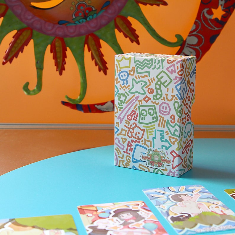 May include: A white rectangular box covered in colorful doodle art, including shapes, faces, and symbols. The box sits on a light blue surface, with illustrated cards in the foreground and a decorative orange background.