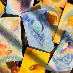 May include: A collection of tarot cards with colorful illustrations and text. The cards feature images of birds, flowers, and abstract designs in shades of blue, yellow, and orange. Several dice are scattered among the cards.