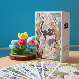May include: A white box featuring a colorful illustration with a black cat, a rabbit, and a person. The box has the text "The KING'S Theater". A small bouquet of colorful pipe cleaner flowers sits on a wooden base. Several cards are in the foreground.
