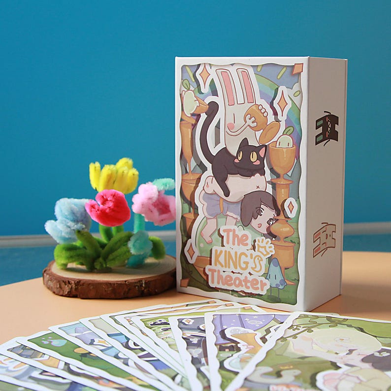 May include: A white box featuring a colorful illustration with a black cat, a rabbit, and a person. The box has the text "The KING'S Theater". A small bouquet of colorful pipe cleaner flowers sits on a wooden base. Several cards are in the foreground.