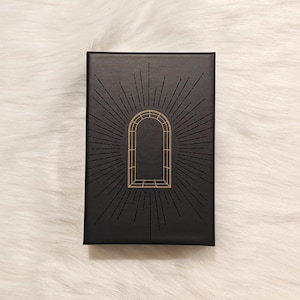 Könnte beinhalten: Eine rechteckige schwarze Schachtel mit einem goldenen geometrischen Bogendesign und strahlenförmigen Linien. Die Schachtel befindet sich vor einem weißen, flauschigen Hintergrund. Das Design ist auf der Schachtel zentriert und erzeugt ein symmetrisches und elegantes Aussehen.