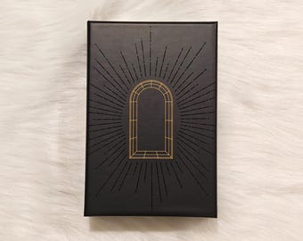 May include: A rectangular black box with a gold geometric arch design and radiating lines. The box is set against a white, fluffy background. The design is centered on the box, creating a symmetrical and elegant appearance.