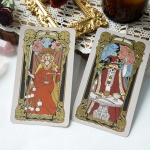 May include: Two tarot cards with gold trim and ornate designs. The card on the left features a woman in a red robe holding a sword and scales. The card on the right features a man in a red robe holding a sword and a cup.