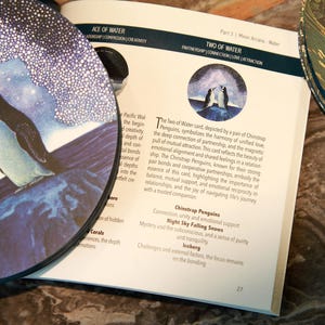 May include: A tarot card deck with a circular card featuring two penguins on a blue and white background. An open book displays text about the "Two of Water" card, with a decorative plate in the background.