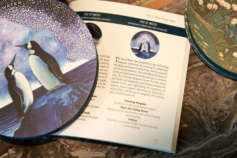 May include: A tarot card deck with a circular card featuring two penguins on a blue and white background. An open book displays text about the "Two of Water" card, with a decorative plate in the background.