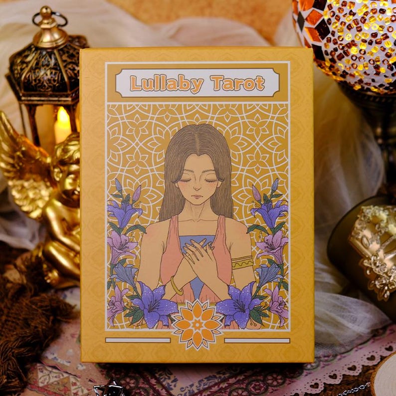 K&ouml;nnte beinhalten: Eine Tarotkartenbox mit dem Titel "Lullaby Tarot". Die Box zeigt eine Illustration einer Frau mit langen braunen Haaren, die ein rosa-blaues Outfit tr&auml;gt, umgeben von lila und blauen Blumen. Der Hintergrund ist goldgelb.
