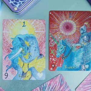 May include: Two rectangular tarot cards featuring illustrations of a blue unicorn and a knight on horseback. The left card shows a blue unicorn with a yellow mane and the number 9, while the right card depicts a knight on a blue horse with the text Rider.