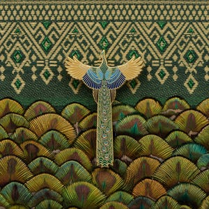 May include: A gold-colored peacock pin with blue and green accents. The pin is set against a background of green and gold patterned fabric and a bed of peacock feathers.