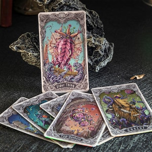 May include: A collection of rectangular trading cards featuring intricate, ornate borders and colorful illustrations of mythical creatures. One card stands upright, labeled Elder Thing, while others are fanned out, showing Hastur and Yog-Sothoth.