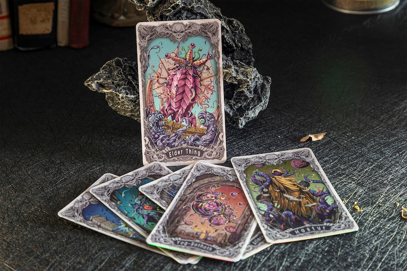 May include: A collection of rectangular trading cards featuring intricate, ornate borders and colorful illustrations of mythical creatures. One card stands upright, labeled Elder Thing, while others are fanned out, showing Hastur and Yog-Sothoth.