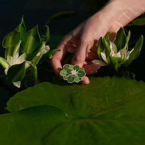 May include: A green and white enamel pin featuring a stylized water lily with a white flower and green leaves. The pin is held in a hand over a green lily pad.