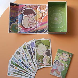 May include: An open, illustrated box with a green interior and a fan of tarot cards. The cards feature colorful illustrations, including a couple embracing and the text "VI-THE LOVERS." A smaller card reads "The King's Theater."