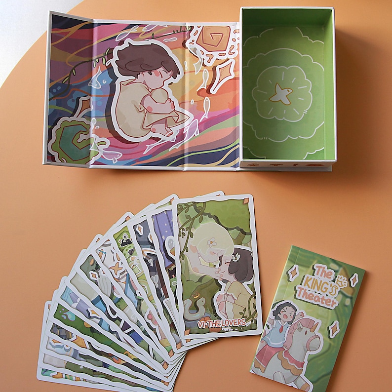 May include: An open, illustrated box with a green interior and a fan of tarot cards. The cards feature colorful illustrations, including a couple embracing and the text "VI-THE LOVERS." A smaller card reads "The King's Theater."