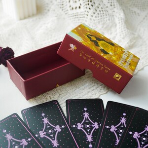 May include: A red box with a gold lid containing a deck of tarot cards. The lid features an illustration of a woman with a crown and a golden background. The text on the lid reads "The Land of Stories Tarot". The cards are black with purple accents and feature a geometric design.