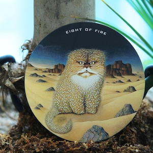 May include: Round artwork featuring a cat in a desert landscape. The cat has a fluffy, spotted coat and a stern expression. The text "EIGHT OF FIRE" is at the top. The background shows mountains and a blue sky.