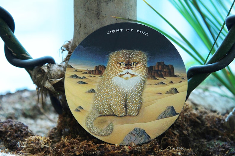May include: Round artwork featuring a cat in a desert landscape. The cat has a fluffy, spotted coat and a stern expression. The text "EIGHT OF FIRE" is at the top. The background shows mountains and a blue sky.