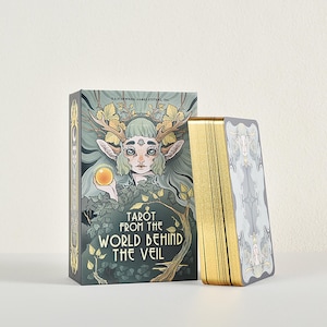 May include: A deck of Tarot from the World Behind the Veil cards with a box featuring a green-haired figure with antlers holding a glowing orb. The box displays the title in white text, and the card edges are finished with a metallic gold foil.