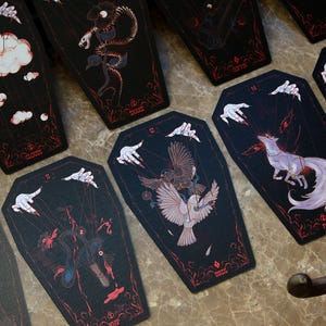 May include: A collection of coffin-shaped tarot cards featuring black backgrounds with red line art and illustrations of hands manipulating strings attached to various subjects including clouds, a snake, birds, and a fox with text labels.