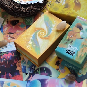 May include: An assortment of tarot cards and boxes with the text "Uno with the Sun". The boxes are yellow, orange, and teal. The cards feature colorful illustrations and are scattered around a small bird's nest with stickers.